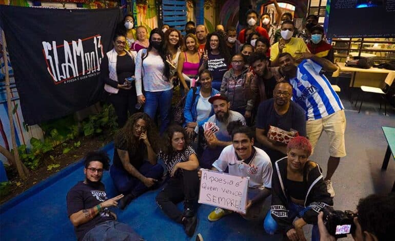 Foto mostra vários participantes do Poesia Falada, ao lado de uma bandeira na cor preta, escrito "Slam do 13"