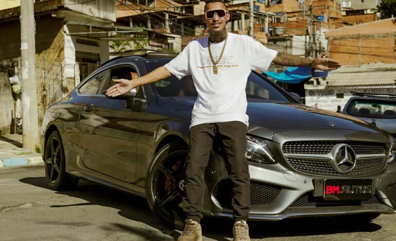 Foto mostra MC Vitinho de camiseta branca, calça preta e tênis de braços abertos em frente a um carro cinza