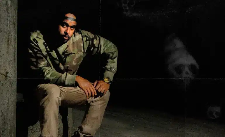 Foto mostra o artista Emcee Canela sentado, usando boné, camisa de manga comprida verde camuflada e calças marrom.
