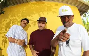 Foto mostra os integrantes do grupo Conteúdo Paralelo, olhando para a camera os 3 artistas estão em frente a um muro amarelo, todos usam bonés, calças jeans e camiseta branca