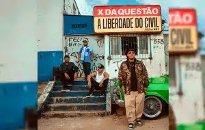 Quatro homens posam em frente a um prédio desgastado com grafites e escadas de concreto. Dois estão sentados nos degraus e dois em pé. Ao fundo, há uma placa com os dizeres “X da Questão – A Liberdade do Civil Volume #01” e um carro antigo verde estacionado à direita. O ambiente remete à cultura de rua e ao estilo urbano.